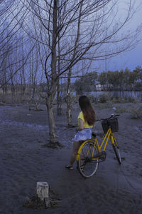 Rear view of young woman with bicycle standing on field against sky at dusk