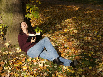 Full length of woman laughing while holding book by tree at park during autumn