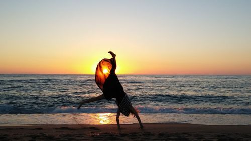 Teenage girl doing cartwheel at beach against sky during sunset