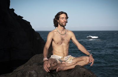 Full length of shirtless man meditating while sitting on rock against sea