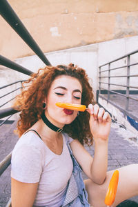 Close-up woman puckering while having orange popsicles on footpath