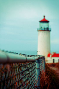 Chainlink fence on field towards lighthouse against blue sky