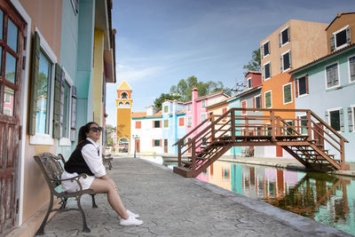 Woman sitting on chair by canal against buildings