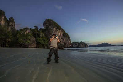 Man photographing while standing on beach against sky during sunset