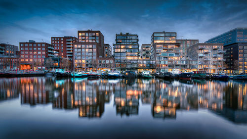 Illuminated buildings by river against sky in city at dusk