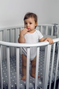 One year old child in white clothes standing in a white round bed in his nursery