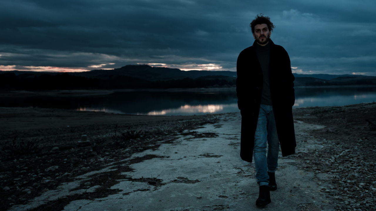 YOUNG MAN STANDING IN LAKE AGAINST SKY DURING SUNSET