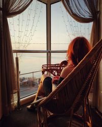 Woman looking through window while sitting on seat at home during sunset