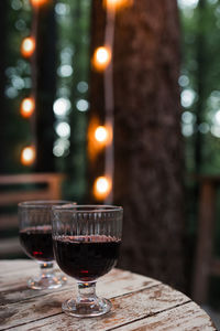 Two glasses of red wine standing on the wooden table with blurred trees and lights 