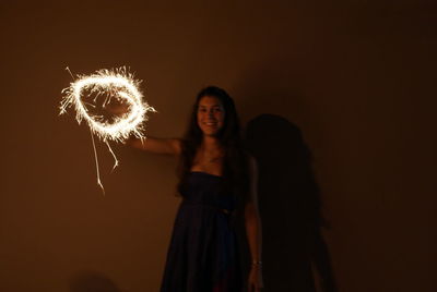 Portrait of woman holding illuminated sparkler against wall at night