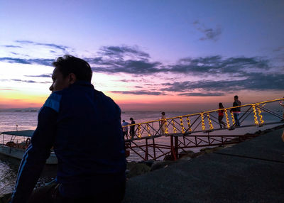 Man standing on railing against sea during sunset