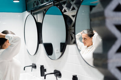 Side view of female worker in elegant wear making ponytail and looking at mirror while standing in restroom with modern design