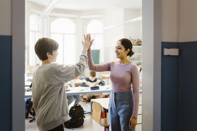 Smiling female teacher giving high-five to schoolboy standing in classroom