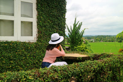 Rear view of woman with hat sitting by window outdoors