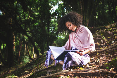 Young woman with a curly hair sitting on a floor with tree roots in a park reading a book