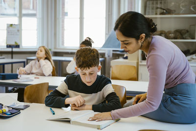 Female teacher helping schoolboy reading in book while sitting on desk in classroom