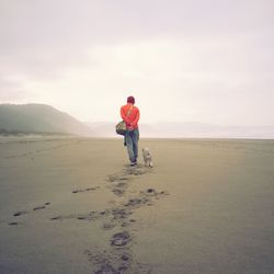 Rear view of man with dog walking on sand against sky