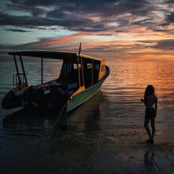 Rear view of young woman standing by boat at shore during sunset