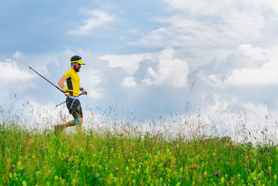 Young man with beard runs downhill in the green grass while practicing nordic walking