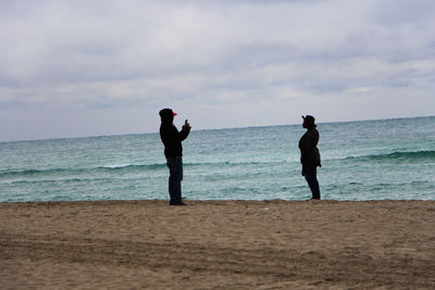 Side view of man photographing woman while standing on shore at beach against cloudy sky