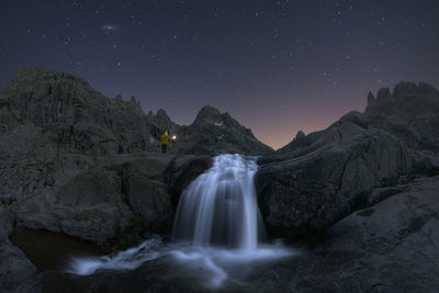 Traveler admiring cascade with foam on rough mount against pond under starry sky at dusk