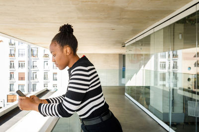 Side view of focused african american female in casual wear text messaging on cellphone while standing on balcony with glass wall