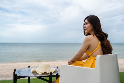 Smiling woman looking away while sitting against sky at beach