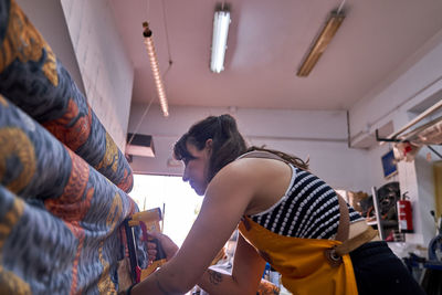 Side view of focused adult female artisan with long dark hair in casual clothes and apron using staple gun while fixing ornamental tapestry on furniture during work in studio