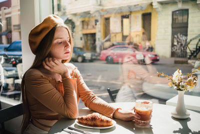 French female in beret sitting at table in cafe with aromatic glass of coffee and freshly baked croissant