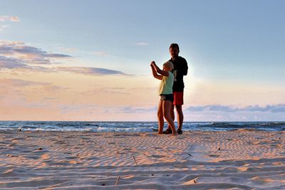 Portrait of father and daughter standing at beach against sky during sunset