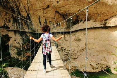 Rear view of woman walking on footbridge leading towards rock formations