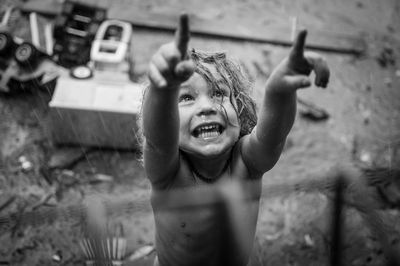 High angle view of happy shirtless boy with arms raised standing outdoors during rainfall