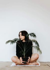 Young woman holding potted plant while sitting on floor against wall