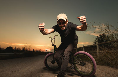 Man with clenched fist sitting on bicycle against sky during sunset