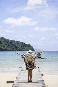 Rear view of woman with arms outstretched standing on pier at beach against sky