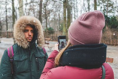 Rear view of woman photographing man while standing at park during winter