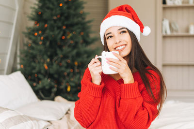 Young asian woman in red sweater and santa hat with cup of cocoa on bed in room with christmas tree