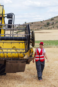Female worker in uniform examining combine harvester while working in agricultural field in countryside on summer day