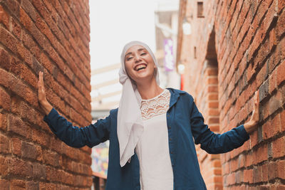 Portrait of cheerful young woman standing by brick wall