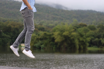 Low section of man in mid-air with hands in pockets over lake against tree mountain