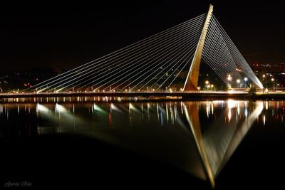 Illuminated suspension bridge over river against sky at night