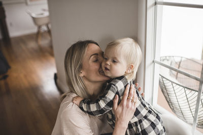 Happy mother kissing daughter by window at home