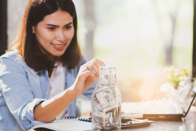 Smiling young woman putting paper currency in jar on table