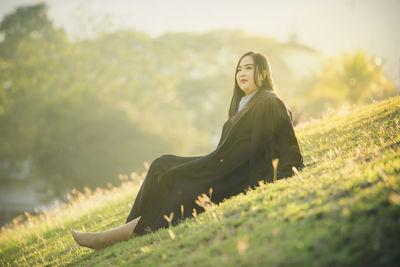Thoughtful woman sitting in graduation gown on grassy field