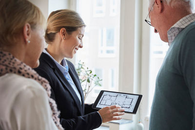 Female real estate agent showing new house blueprint on digital tablet to senior couple at home