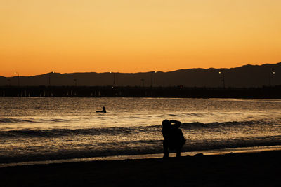 Silhouette man photographing friend at beach against clear sky during sunset