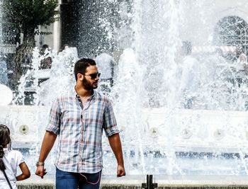 Fashionable man wearing sunglasses while looking away against fountain in city