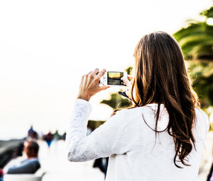 Rear view of woman photographing from mobile phone against clear sky