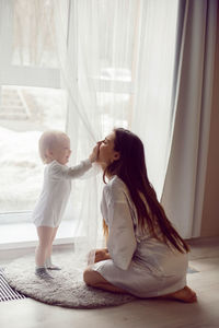 Mother in a white robe sits with a child a blonde daughter at a large window of the house person