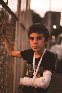 Portrait of boy with broken hand standing by fence during sunset